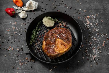 Ossobuco zeytinyağı ve bitkilerle marine edilmiş. Siyah bir demir tavanın üzerinde yatıyorlar. Tabaklar karanlık bir taşın üzerinde duruyor, baharatlar ve bitkiler etrafa saçılmış..