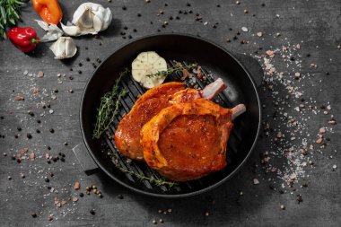 Paprika salamurasında kemikte domuz eti. Siyah dökme demir bir tavanın üzerinde duruyor. Tabaklar karanlık bir taşın üzerinde duruyor, baharatlar ve bitkiler etrafa saçılmış..