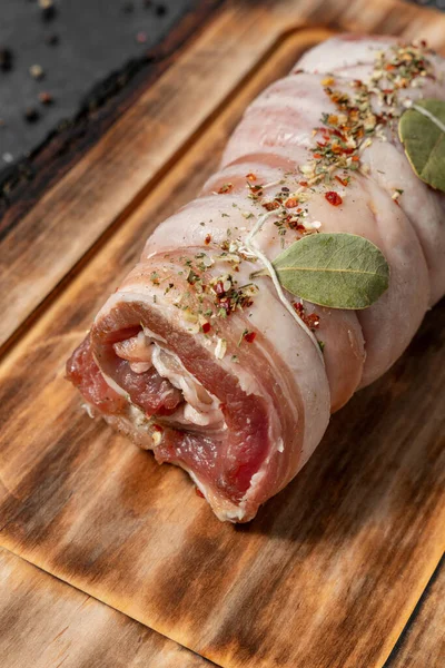 Baharatlı otlar ve defne yaprağıyla domuz eti rulosu. Açık tahta bir tahtanın üzerinde duruyor. Tabaklar karanlık bir taşın üzerinde duruyor, baharatlar ve bitkiler etrafa saçılmış..