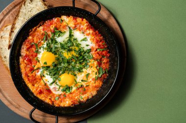 Shakshuka üç yumurta ve doğranmış yeşilliklerle birlikte metal bir tencerede. Yakınlarda iki kadeh beyaz ekmek var. Ahşap bir tezgahta, kağıt renkli arka planda yemek..