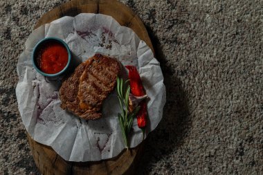 Domuz bifteği, tahta bir tahtanın üzerindeki beyaz parşömenin üzerinde duruyor. Yakınlarda bir kase domates sosu ve biberiye var..