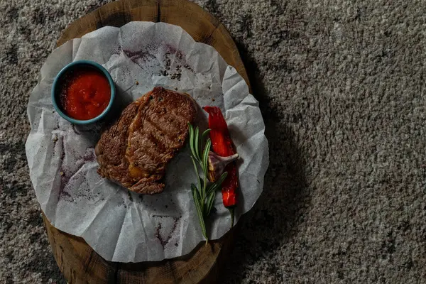 Domuz bifteği, tahta bir tahtanın üzerindeki beyaz parşömenin üzerinde duruyor. Yakınlarda bir kase domates sosu ve biberiye var..