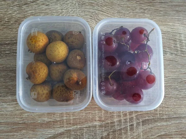 Complementary food supplies for children, in the form of fruit. Two small plastic boxes containing grapes in one box and longans in the other. Contains lots of vitamins and antioxidants.