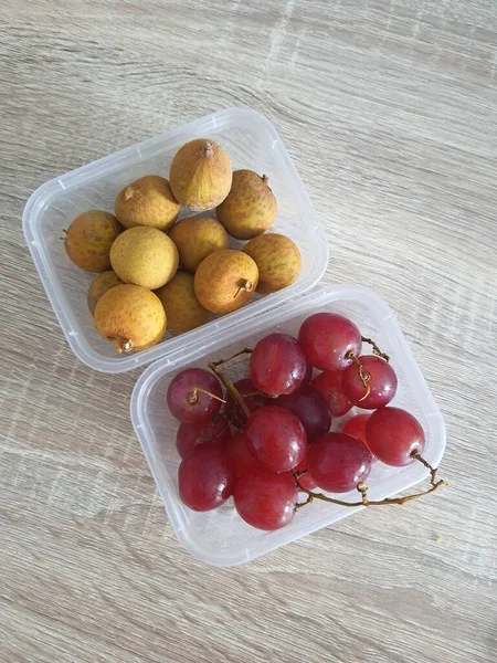 Complementary food supplies for children, in the form of fruit. Two small plastic boxes containing grapes in one box and longans in the other. Contains lots of vitamins and antioxidants.