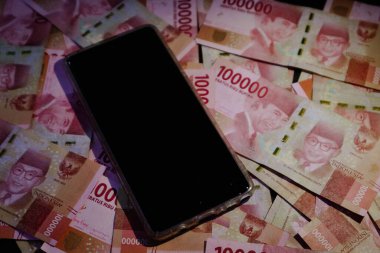 Yüzlerce bin rupia banknotunun üzerinde siyah ekranı olan bir cep telefonu. Şablon geçmişiniz ya da teknoloji, iş ve finans ile ilgili haberler için uygundur..