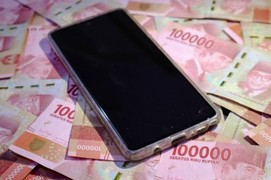 Yüzlerce bin rupia banknotunun üzerinde siyah ekranı olan bir cep telefonu. Şablon geçmişiniz ya da teknoloji, iş ve finans ile ilgili haberler için uygundur..