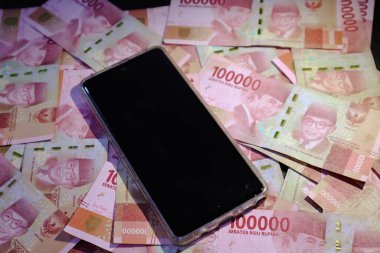 Yüzlerce bin rupia banknotunun üzerinde siyah ekranı olan bir cep telefonu. Şablon geçmişiniz ya da teknoloji, iş ve finans ile ilgili haberler için uygundur..