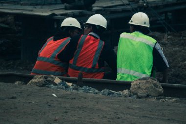 Cakarta, Endonezya Temmuz 2022. Üç inşaat işçisi işten sonra dinlenmek için inşaat sahasının kenarında oturuyorlar, hala kask ve yelek giyiyorlar..