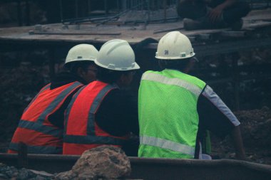 Cakarta, Endonezya Temmuz 2022. Üç inşaat işçisi işten sonra dinlenmek için inşaat sahasının kenarında oturuyorlar, hala kask ve yelek giyiyorlar..