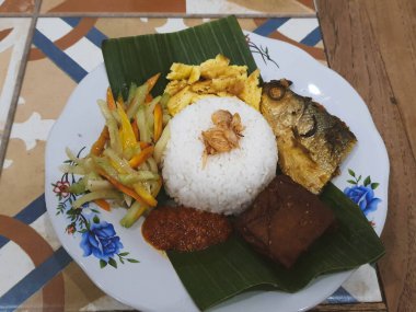 Nasi Kucing 'in bir kısmı kızarmış sütçü balığı başlıklı, tofu, muz yapraklı bir tabakta servis ediliyor. Pirincin üstüne kızarmış soğan serpilmiş. Yendiğinde tadı daha lezzetli olsun diye..