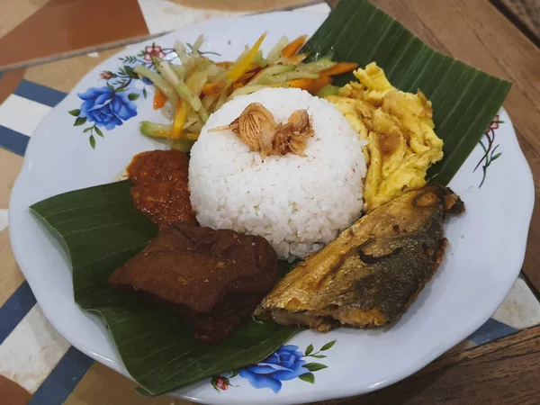 A portion of Nasi Kucing complete with fried milkfish head, tofu, served on a plate with banana leaves. On top of the rice sprinkled with fried onions to make it taste more delicious when eaten.