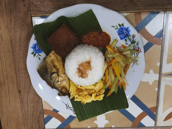 A portion of Nasi Kucing complete with fried milkfish head, tofu, served on a plate with banana leaves. On top of the rice sprinkled with fried onions to make it taste more delicious when eaten.