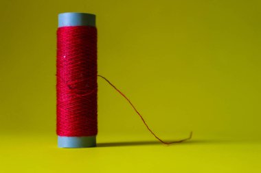 Isolated photo of a roll or a spool of red thread above and in front of a yellow background. This thread is a bit loose from the reel.