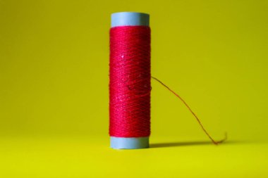 Isolated photo of a roll or a spool of red thread above and in front of a yellow background. This thread is a bit loose from the reel.