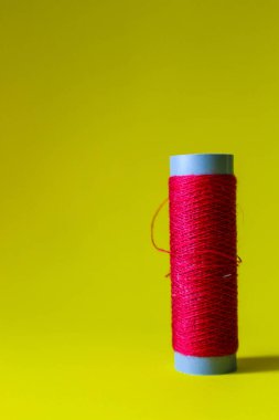 Isolated photo of a roll or a spool of red thread above and in front of a yellow background. This thread is a bit loose from the reel.