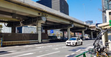 Tokyo, Japan in July 2019. A white sedan car is driving down a quiet Tokyo street. This small road is next to a bridge with a steel box girder structure, and the pier head structure has steel ribs.