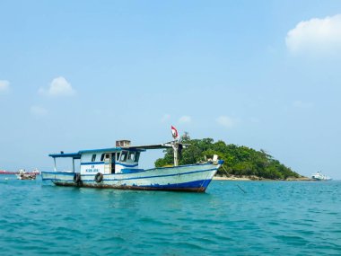 A small island in the port of Merak, Cilegon, Banten. You can see the blue sea, a small island with green trees and a fishing boat around.