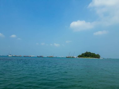 A small island in the port of Merak, Cilegon, Banten. You can see the blue sea, a small island with green trees and a fishing boat around.