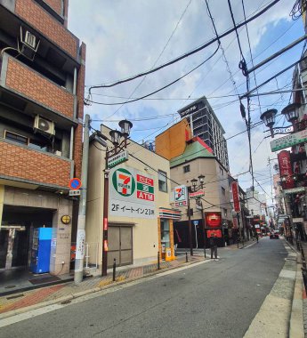 Osaka, Japonya 05 Ekim 2023 'te dar bir Japon sokağı solunda binalar ve kablolar bulunan bir Seven Eleven mağazası gösteriyor. Cadde nispeten sessiz ve birkaç kişi uzaktan görülüyor. Genel ruh hali sakin ve hafifçe