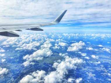 Açık mavi gökyüzünün altında uçsuz bucaksız, tüylü, beyaz bulutları gösteren bir uçak penceresinden çarpıcı bir hava manzarası. Uçak kanadı kısmen ön planda görülebilir, yükseklik ve seyahat hissi katar. Genel havası sakin ve güzel.