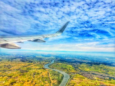 Bir uçak penceresinden nefes kesici bir hava manzarası aşağıdaki tarlaların bir kısmını kesen dolambaçlı bir nehri gösteriyor. Pofuduk bulutlarla dolu canlı mavi gökyüzü manzaranın toprak tonlarıyla güzel bir tezat oluşturuyor. Uçak kanadı...
