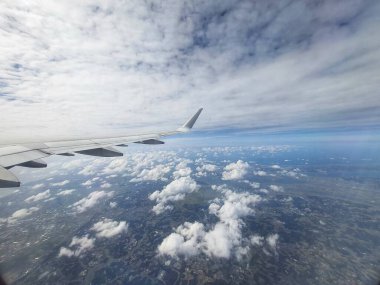 Bir uçak penceresinden yapılan hava görüntüsü, alttaki yamaca yayılmış geniş, tüylü beyaz bulutları gösteriyor. Uçak kanadı kısmen ön planda görülebiliyor ve sahneye bir ölçek ve perspektif katıyor. Genel olarak. 
