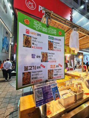 Seul, Güney Kore, 12 Kasım 2024. Myeongdong 'da. Hareketli bir Kore gece marketi sahnesinde renkli bir hotteok standı, menüsü çeşitli tatlı ve lezzetli dolguları sergiliyor. Sıcak, davetkar, lezzetli.!