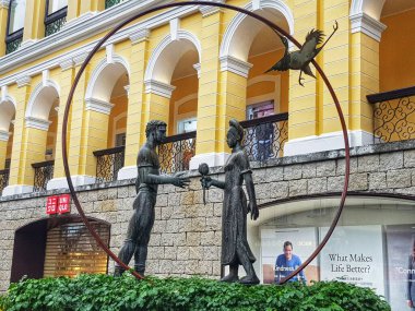 Santo Antonio, Macau 14 Kasım 2024. İki bronz figür, canlı sarı bir binaya karşı dizilmiş bir çiçeği zarifçe değiş tokuş ediyor. Yuvarlak bir çerçeve ve uçan bir kuş tarafından geliştirilmiş sakin ve sanatsal bir sahne..