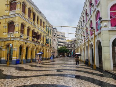Largo do Senado, Macau 14 Kasım 2024. Macau 'nun Senado Meydanı. Yağmurlu bir gökyüzünün altında büyüleyici bir Macau sokak sahnesi sergileniyor. Sarı koloni binaları, altın kemerli bir yapının altında şemsiyeli yayaların dolaştığı dalgalı bir kaldırım taşı caddesi hizasında., 