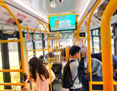 Macau, Çin 14 Kasım 2024. Hareketli bir şehir otobüsü sahnesi. Yolcular telefonlarını kullanır, parlak sarı tırabzanların yanında dururlar. Dijital ekran, başınızın üstünde dinamik bir atmosfer yaratarak bilgi görüntüler.