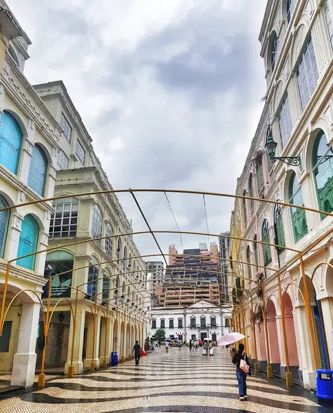 Largo do Senado, Macau 14 Kasım 2024. Macau 'nun Senado Meydanı. Büyüleyici bir Macau oyun salonu yağmurlu bir gökyüzünün altında açılıyor. Pastel renkli binalar, başınızın üzerinde altın kemerler ve pembe şemsiyeli bir insan huzurlu, nostaljik bir atmosfer yaratır..