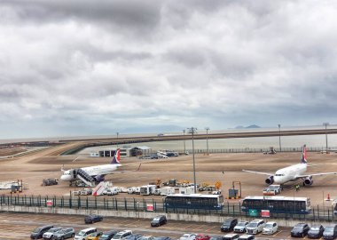 Macau, Çin 14 Kasım 2024. Bulutlu gökyüzü hareketli bir havaalanı pistinin üzerinde asılı. İki uçak park halinde, yer destek araçları ve dolu bir park yeri ile çevrili. Barışçıl ama operasyonel bir atmosfer.