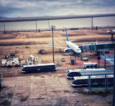 Macau, Çin 14 Kasım 2024. Bulutlu bir gökyüzünün altında sakin bir havaalanı sahnesi sergileniyor. Bir uçak jet köprüsüne bağlı, yer ekibi çalışıyor, otobüsler bekliyor. Barışçıl ama işlevsel.