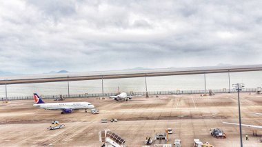 Macau, Çin 14 Kasım 2024. Sakin bir havaalanı sahnesi, bulutlu bir gökyüzünün altında sakin bir denize bakan engin bir pistte park etmiş iki uçağın görüntüsünü gözler önüne seriyor. Ortam sakin ve huzurlu..