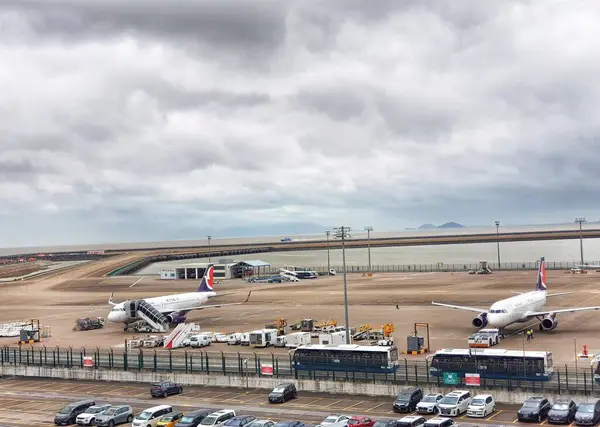 Macau, Çin 14 Kasım 2024. Bulutlu gökyüzü hareketli bir havaalanı pistinin üzerinde asılı. İki uçak park halinde, yer destek araçları ve dolu bir park yeri ile çevrili. Barışçıl ama operasyonel bir atmosfer.