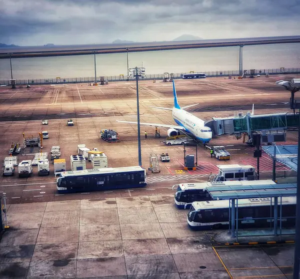 Macau, Çin 14 Kasım 2024. Bulutlu bir gökyüzünün altında sakin bir havaalanı sahnesi sergileniyor. Bir uçak jet köprüsüne bağlı, yer ekibi çalışıyor, otobüsler bekliyor. Barışçıl ama işlevsel.