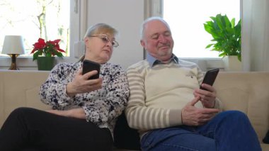 Oturma odasındaki koltukta oturan mutlu yaşlı karı ve koca cep telefonlarıyla internet başvurularını kullanıyor, her biri cep telefonundan gelen haberleri ya da bilgileri paylaşıyor. Yüksek kalite 4k görüntü