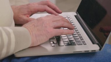 Üst sınıflardan birinin elleri, mutfak tezgahında bulunan dizüstü bilgisayarda yazarken yakın çekim görüntüsü. Doğal ışık odayı doldurarak, yaşlıların günlük yaşamını ve teknolojisini ön plana çıkarıyor. Yüksek kalite