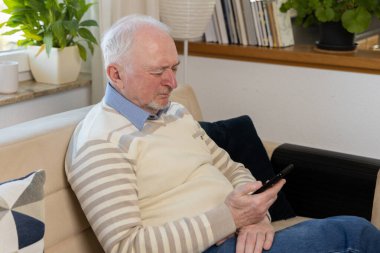 Yaşlı bir adam evdeki kanepede otururken telefonuna bakar. Akıllı telefonunu güncel haberleri, eğlenceyi ve sosyal ağları aktif olarak kullanmak için kullanıyor. Yüksek kalite fotoğraf