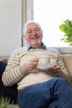 Kanepeyi dinlendirirken elinde kahve tutan mutlu kıdemli adam. Gülümseyen olgun bir adam, modern mutfak oturma odasında rahat rahat otururken kameraya bakarak çay içiyor. Portre.