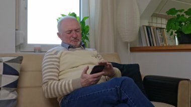 Gri saçlı bir emekli yalnız oturuyor ve telefon kayıtlarında geziniyor. Ekrana odaklanmış durumda. Emeklilerin dijital ortamda eğlenmesi. Yüksek kalite 4k görüntü