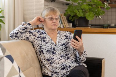 Gri saçlı olgun bir kadın açık kanepede rahat bir şekilde oturuyor akıllı telefonu tutuyor ve ekrana bakıyor. Kadın rahat bir mutfak ortamında rahatlamış görünüyor. Yüksek kalite fotoğraf