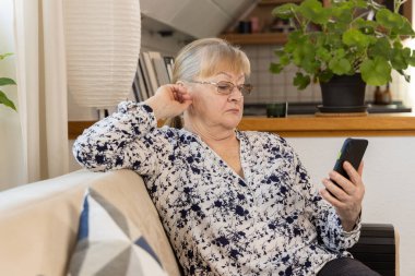 Gri saçlı olgun bir kadın açık kanepede rahat bir şekilde oturuyor akıllı telefonu tutuyor ve ekrana bakıyor. Kadın rahat bir mutfak ortamında rahatlamış görünüyor. Yüksek kalite fotoğraf