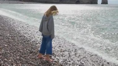 İlkbaharın başında Etretat 'ta kayalık plajda yürüyen günlük kıyafetli genç bir kız. Manş Denizi 'nin sakin suları. güneşli bir gün.