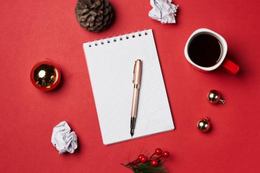 Kırmızı bir yüzey üzerinde bir not defteri, kalem ve noel süslemeleri kahve kupaları ve arka planda çam kozalakları