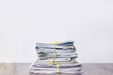 Beyaz bir duvarın önündeki ahşap bir masada, kenarlarında sarı kurdele olan bir yığın gazete.