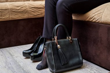 Önünde çantası ve ayakkabılarıyla koltukta oturan bir kadın kameraya bakıyor.