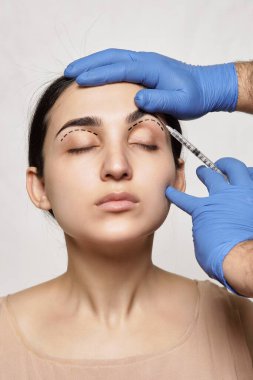 Arka planda profesyonel bir makyajcı tarafından kaşlarını aldıran bir kadın beyaz bir duvardır ve mavi eldivenleri vardır.