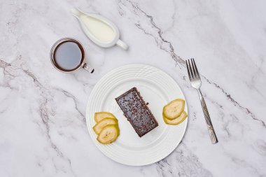 a piece of chocolate cake on a white plate with two apples and a cup of coffee next to the plate