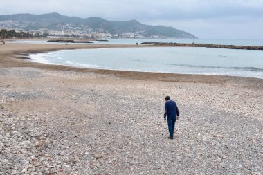 Kasvetli ve yalnız bir adam, boş bir sahilde giyinmiş, deniz kıyısında taş topluyor.